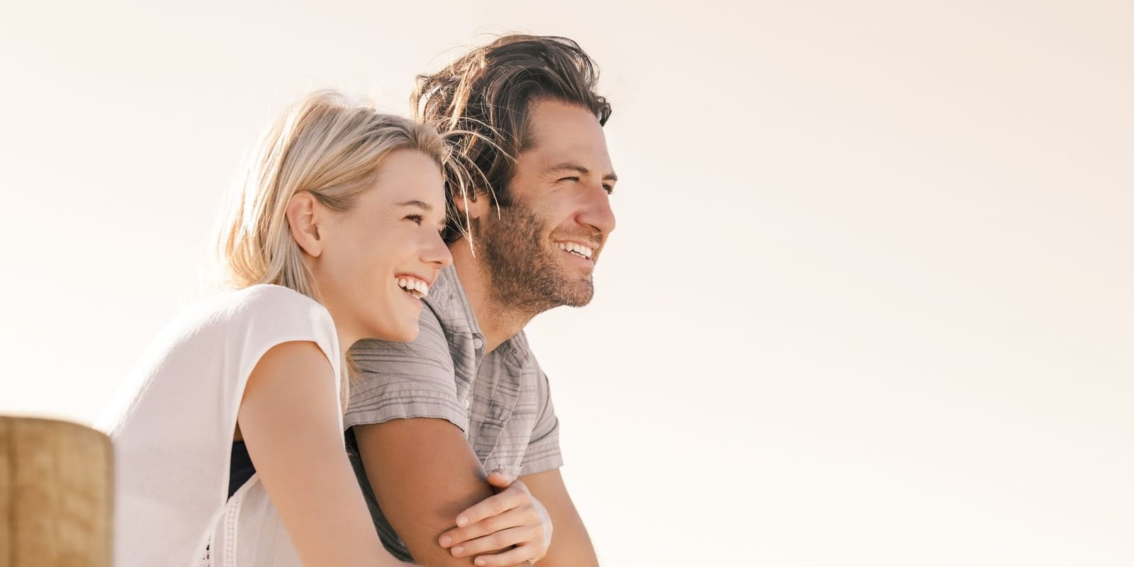A couple outdoors leaning together on a wooden rail, smiling and looking into the distance