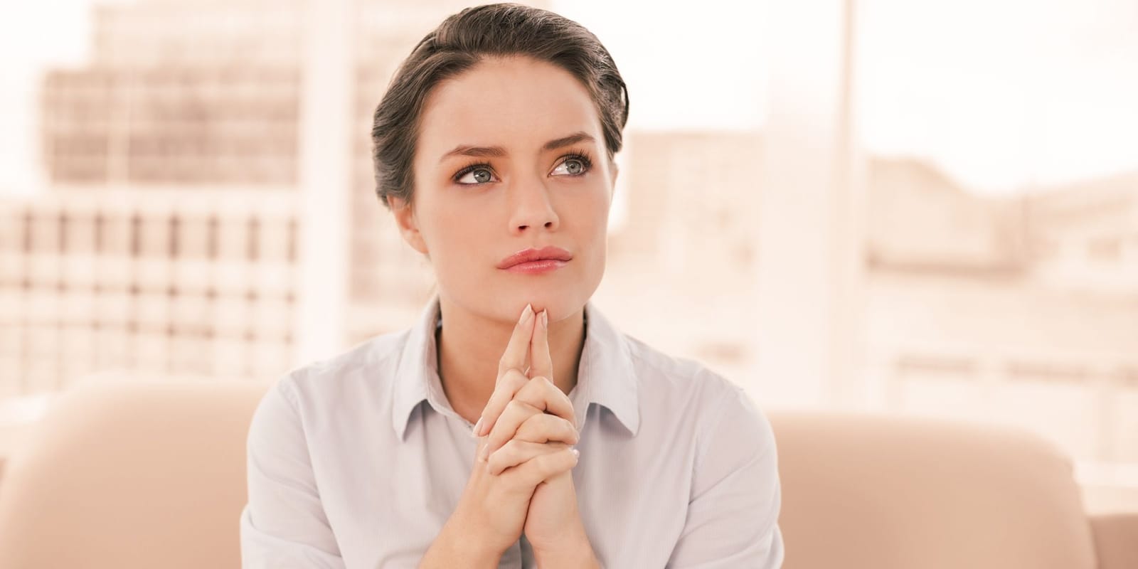A woman sitting on a sofa with her hands clasped beneath her chin, looking thoughtful